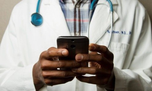 Black hands of someone holding a smart phone with a stathoscope and medical white coat on
